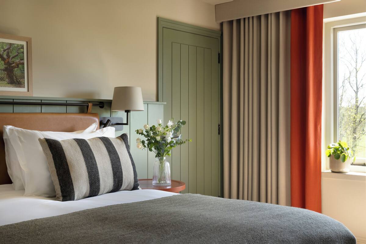 A neatly made bed with pillows, a bedside lamp, a vase with flowers, and a window with curtains.
