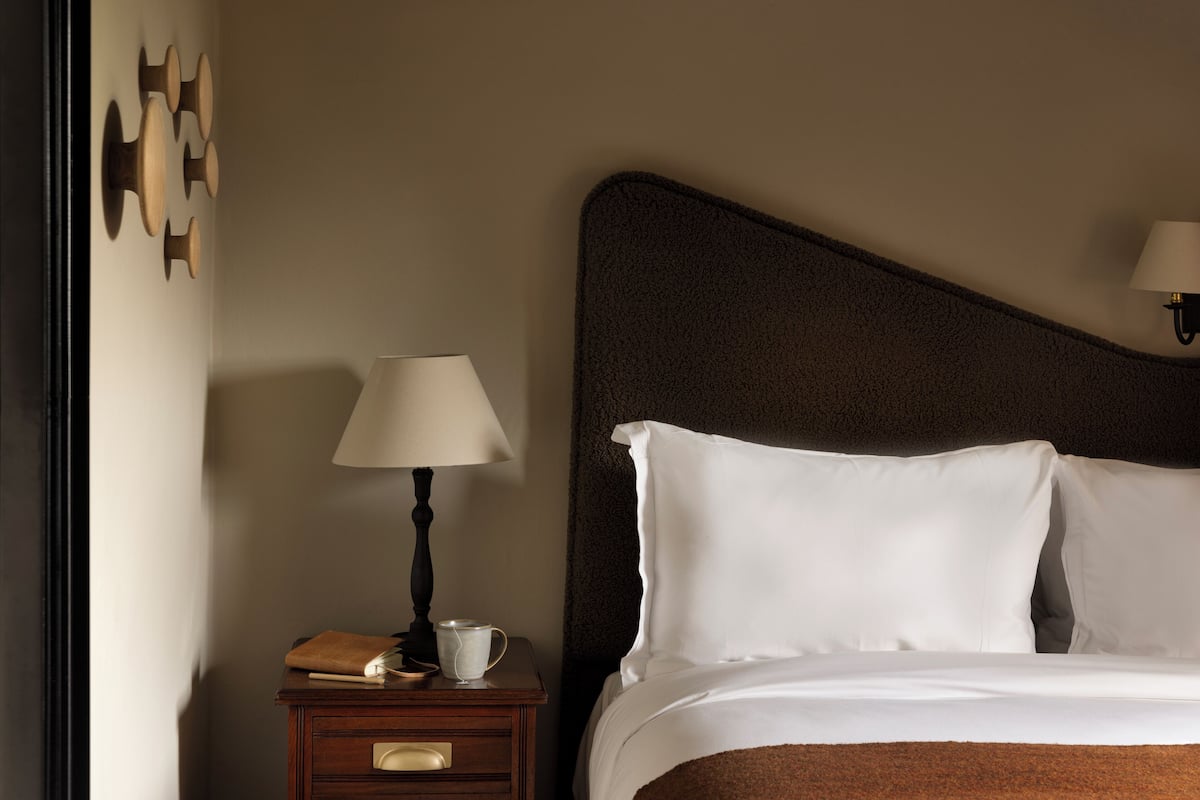 A neatly made bed with white linens, a dark headboard, a bedside table with a lamp, a book, and a cup.