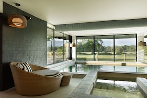 A modern indoor swimming pool area with a wicker chair and cushions, large windows offering a view of the outdoors, and a ceiling light fixture.