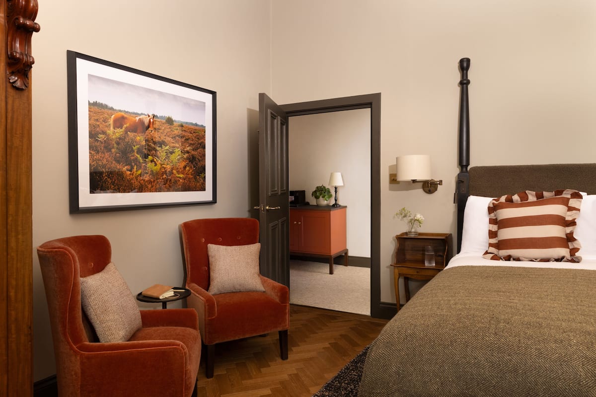 A hotel room with a large framed picture, two red armchairs, a wooden bed, a nightstand with a lamp, and a mirror on the door.