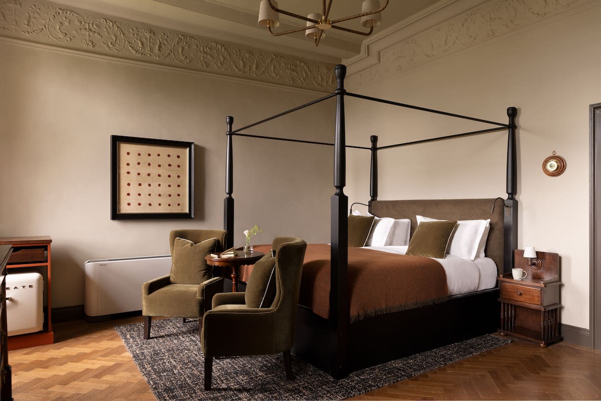 A four-poster bed with a dark headboard, a small bedside table with a lamp, and a chair in a room with a herringbone floor pattern.
