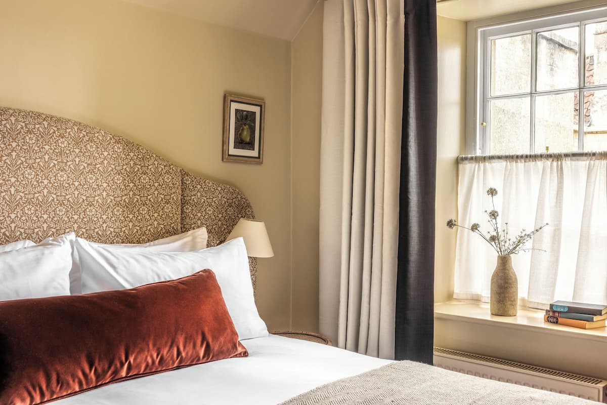 Een net opgemaakt bed met wit en bruin beddengoed, een vaas met bloemen op een vensterbank, en een lijst met een afbeelding aan de muur.