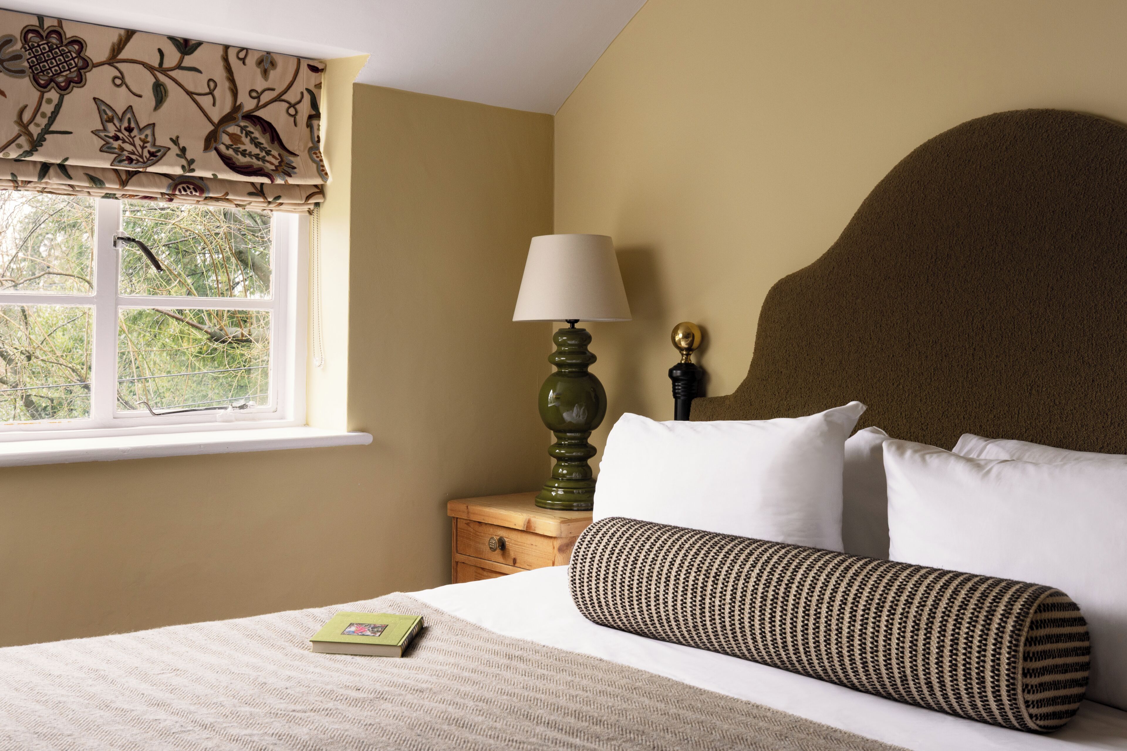 A bedroom with a bed, pillows, a bedside lamp, a nightstand, a window with a view, and a floral-patterned valance.