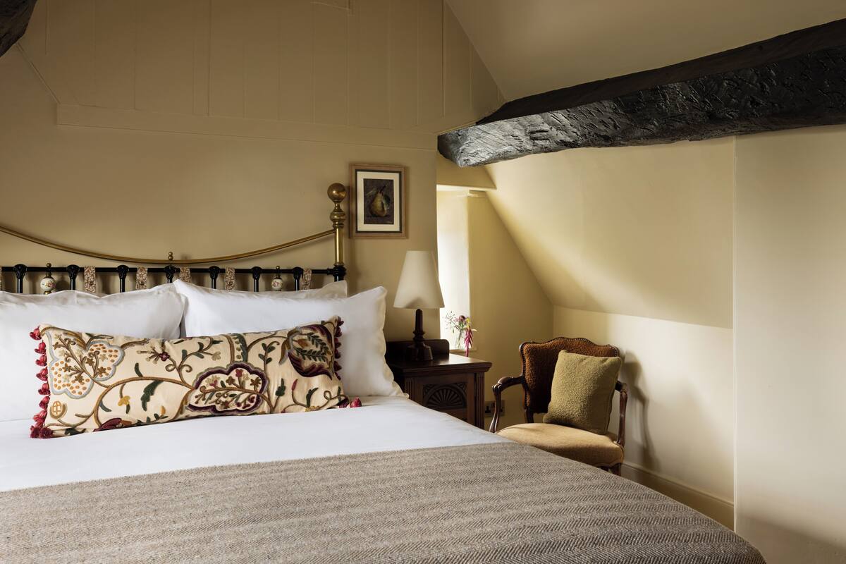 Een keurig opgemaakt bed met een gestreept kussen, een nachtkastje met een lamp, en een stoel in een kamer met een schuine kap en zichtbare balken.