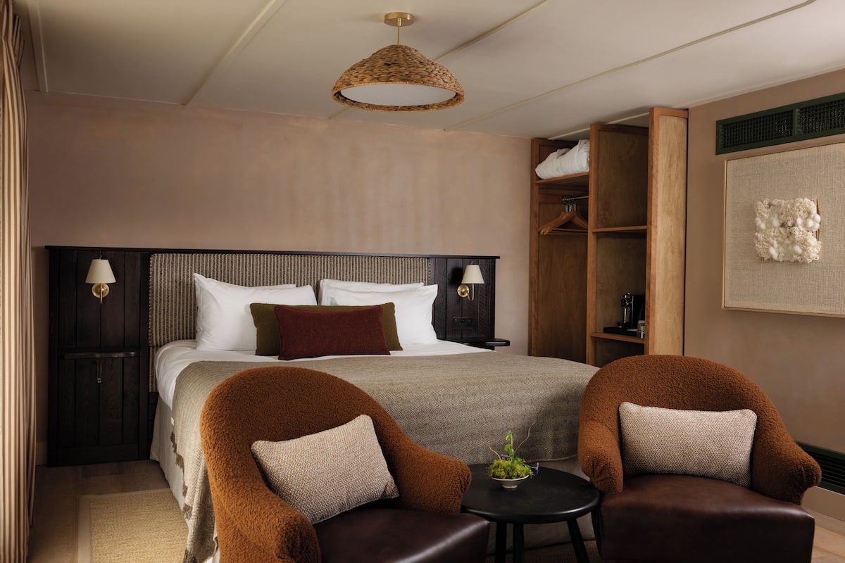 Hotelkamer met een groot bed, twee fauteuils, een klein tafeltje, een geweven plafondlamp, een garderobe en een kunstwerk aan de muur.