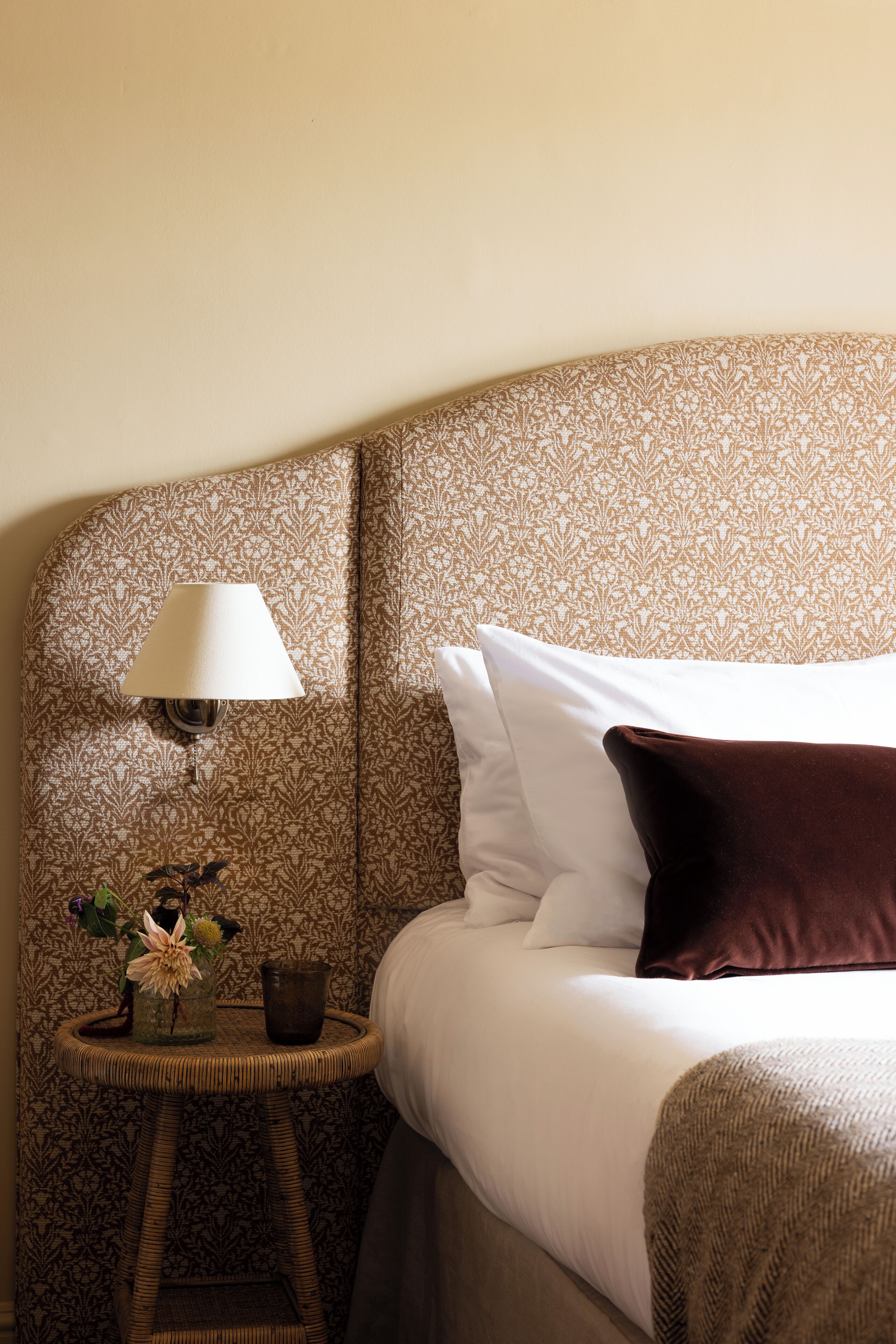 A neatly made bed with white and brown pillows, a small bedside table with a lamp, and a vase with flowers.