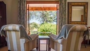Une pièce avec deux fauteuils à rayures placés face à une table, une fenêtre à cadre en bois avec des rideaux et un miroir accroché au mur.