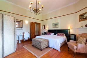 A bedroom with a large bed, a chair, a nightstand, a lamp, a painting, and a mirror.