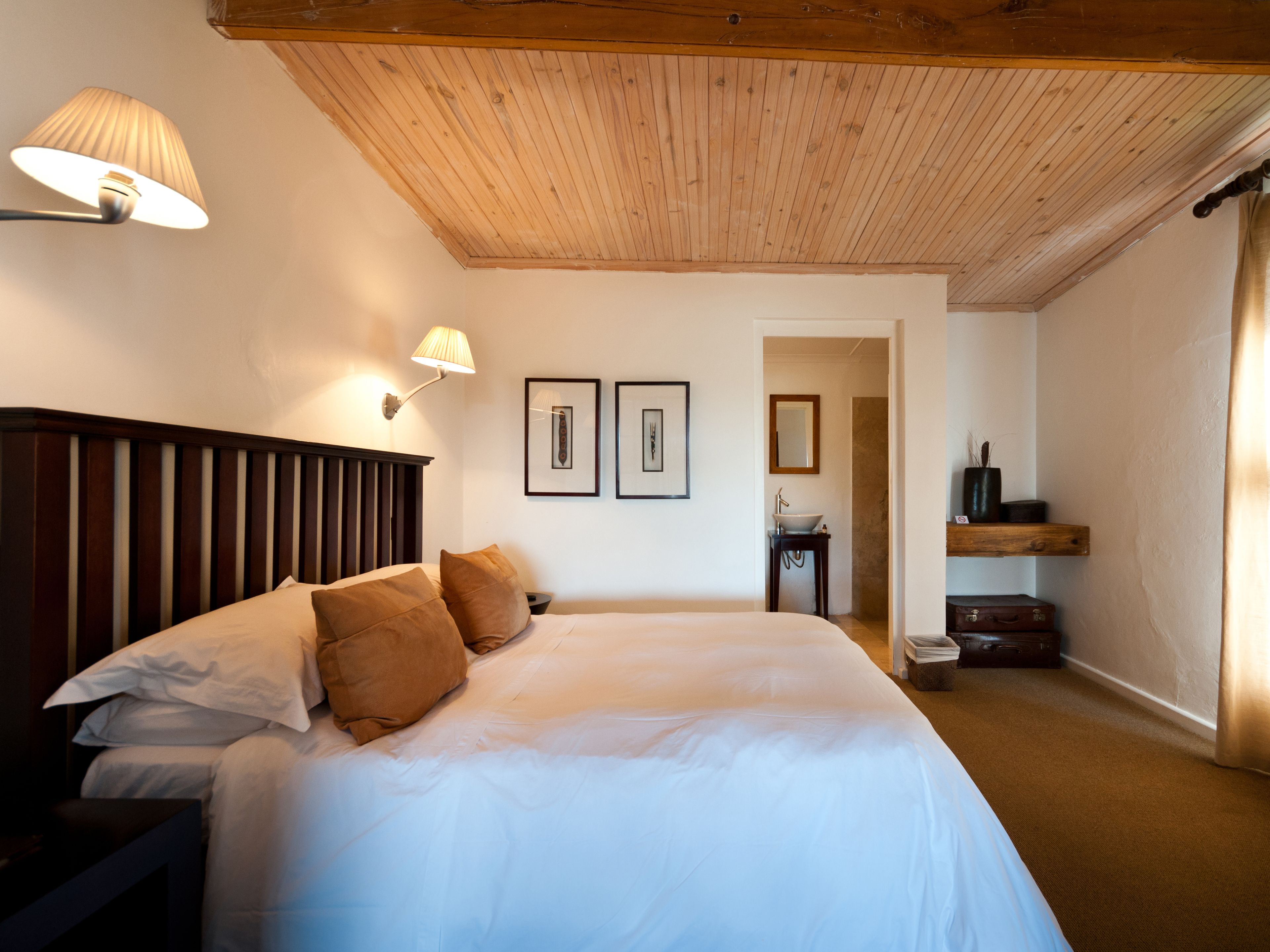 Un dormitorio con techo de madera, una cama con ropa de cama blanca, dos lámparas fijadas a la pared y un cuadro enmarcado.
