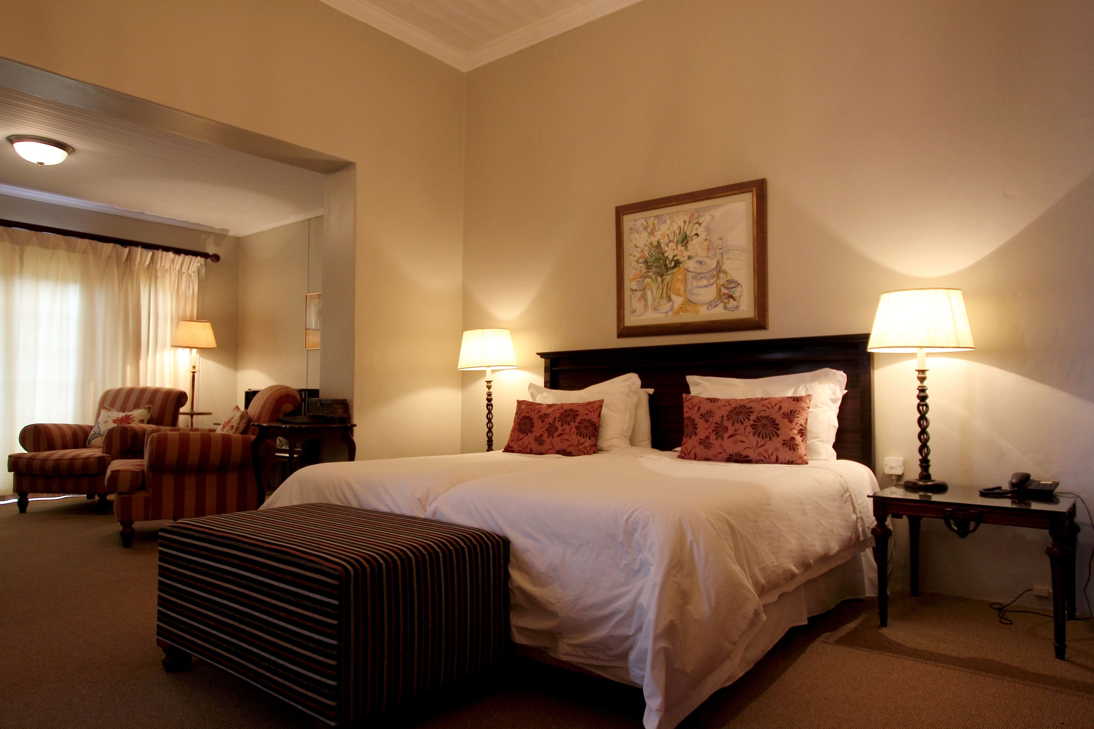 A hotel room with a large bed, two bedside lamps, a headboard with a painting, a chair, a small table, and a striped ottoman.