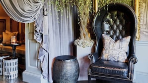 Une chambre luxueuse, avec un fauteuil en cuir foncé, un grand vase rempli de fleurs blanches et un rideau orné de motifs de lierre.