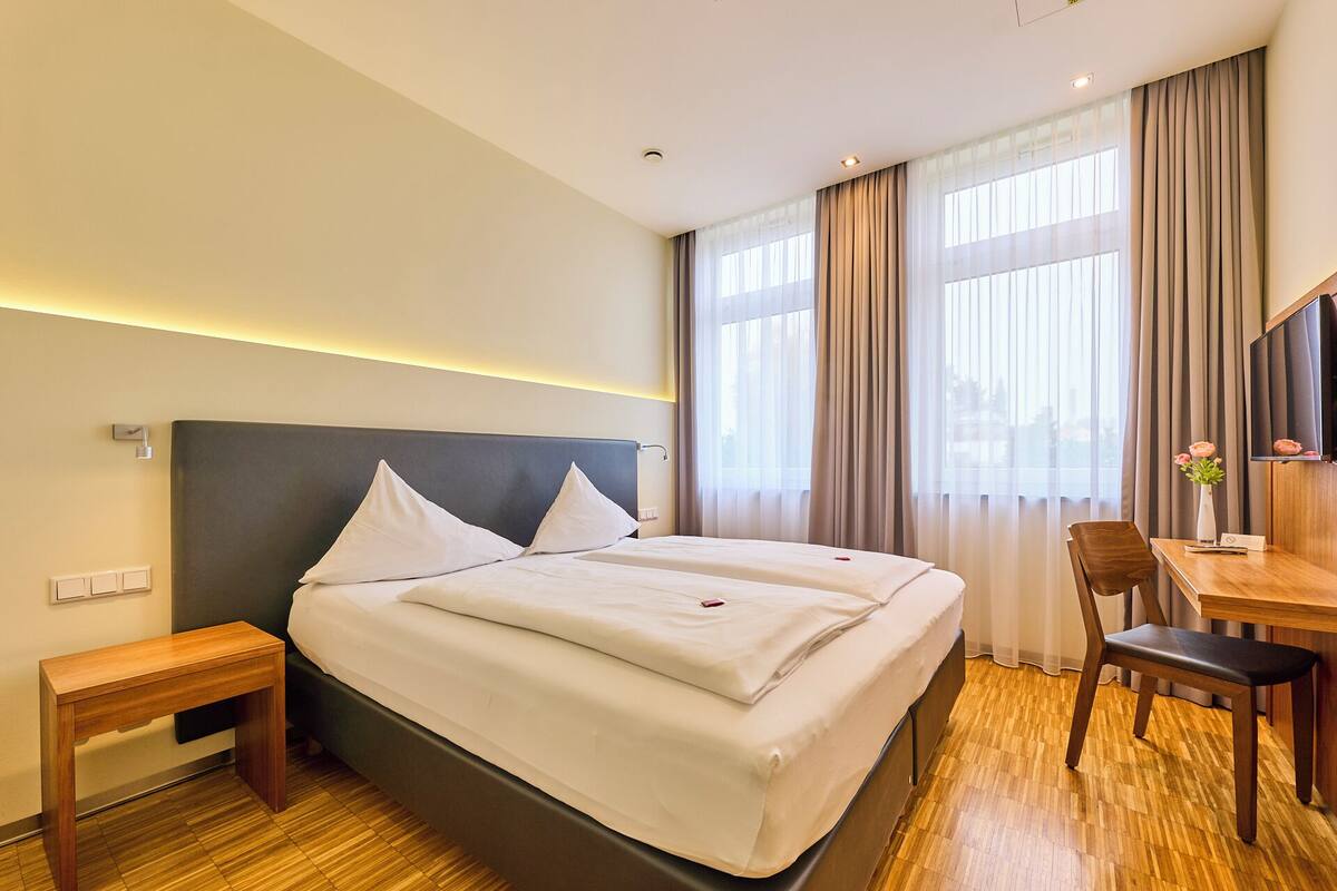 A hotel room with a bed, bedside table, chair, desk, and a TV.