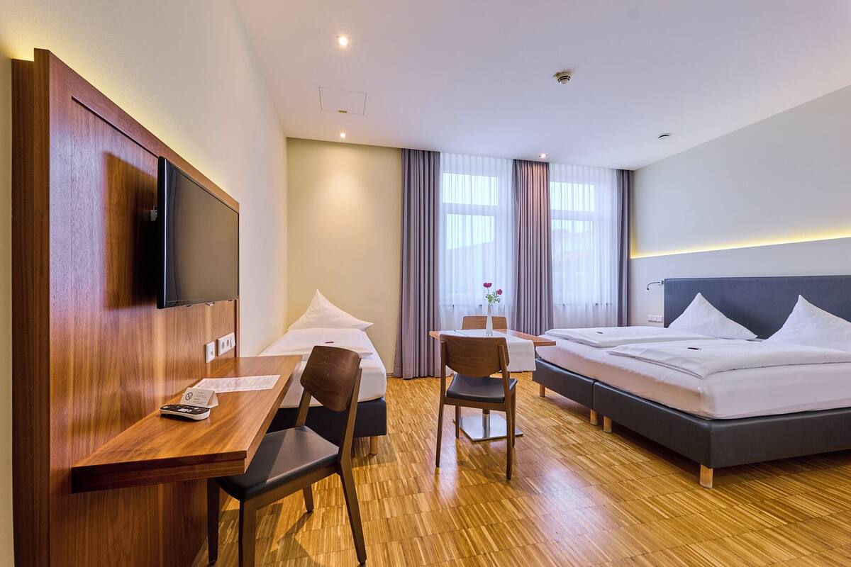 A hotel room with a wooden desk, a bed, a chair, and a TV mounted on the wall.