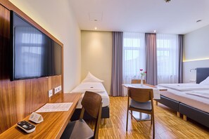 A hotel room with a wooden desk, a chair, a bed, a TV, and a window with curtains. - Hotel Apfelbaum (Erding)