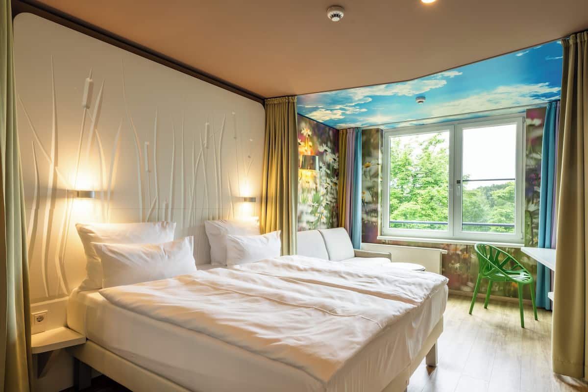 A hotel room with a bed, a chair, a desk, and a window with a view of greenery.