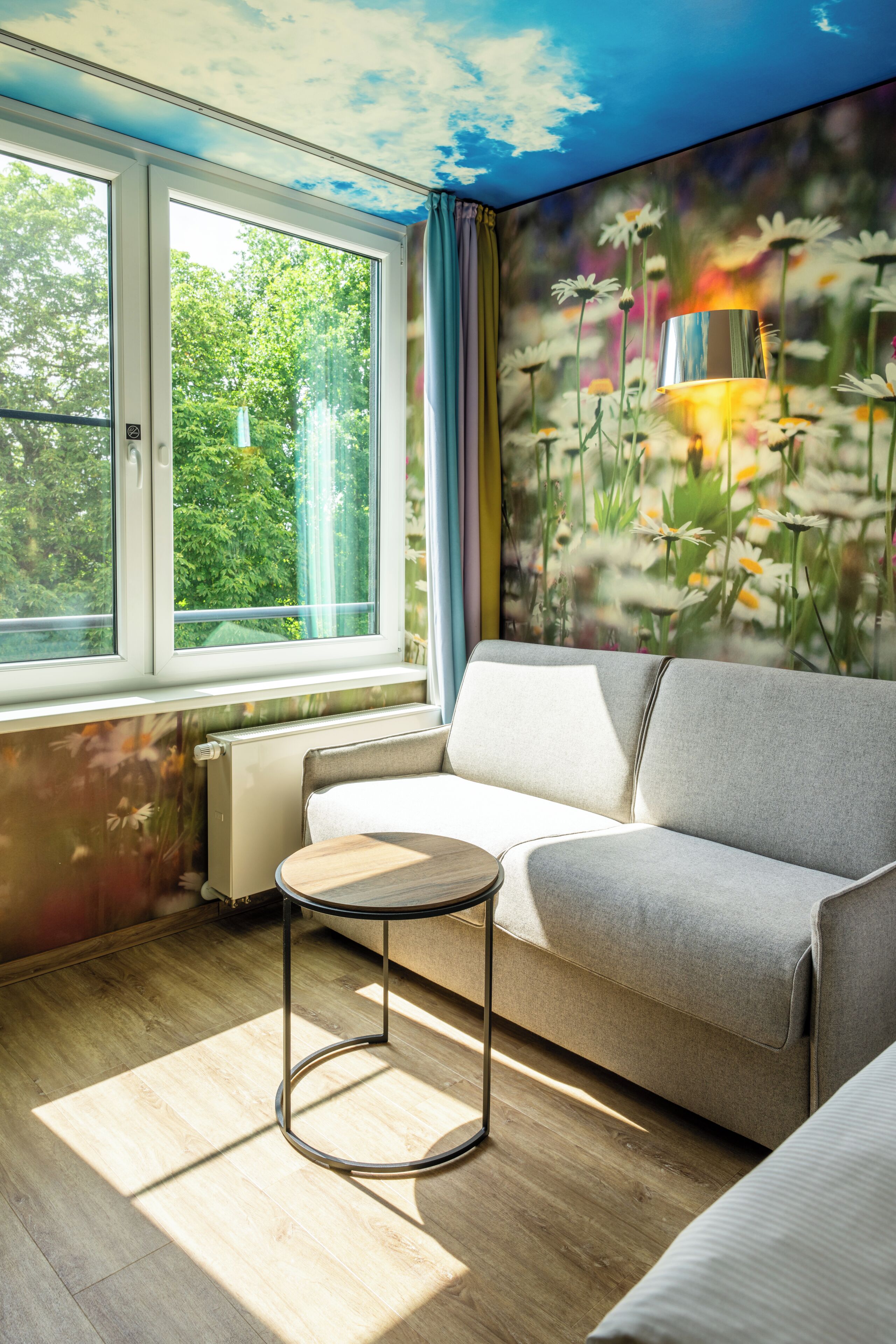 A room with a sofa, a small round table, a radiator, and a window with a view of greenery.