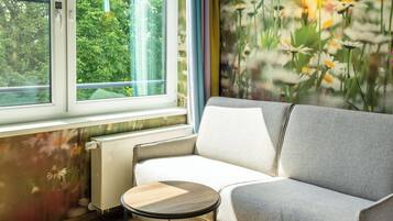 A room with a sofa, a small round table, a radiator, and a window with a view of greenery.