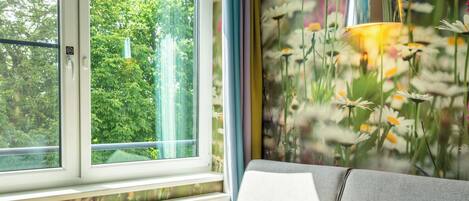 A room with a sofa, a small round table, a radiator, and a window with a view of greenery.