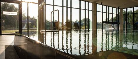 Uma piscina interior com paredes de vidro transparente, uma área banhada pelo sol e uma placa com o texto "SONNENTERRASSE".