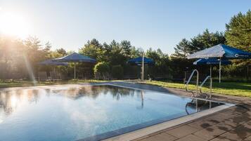 Una piscina con lettini e ombrelloni nelle vicinanze, circondata da alberi e un cielo sereno.