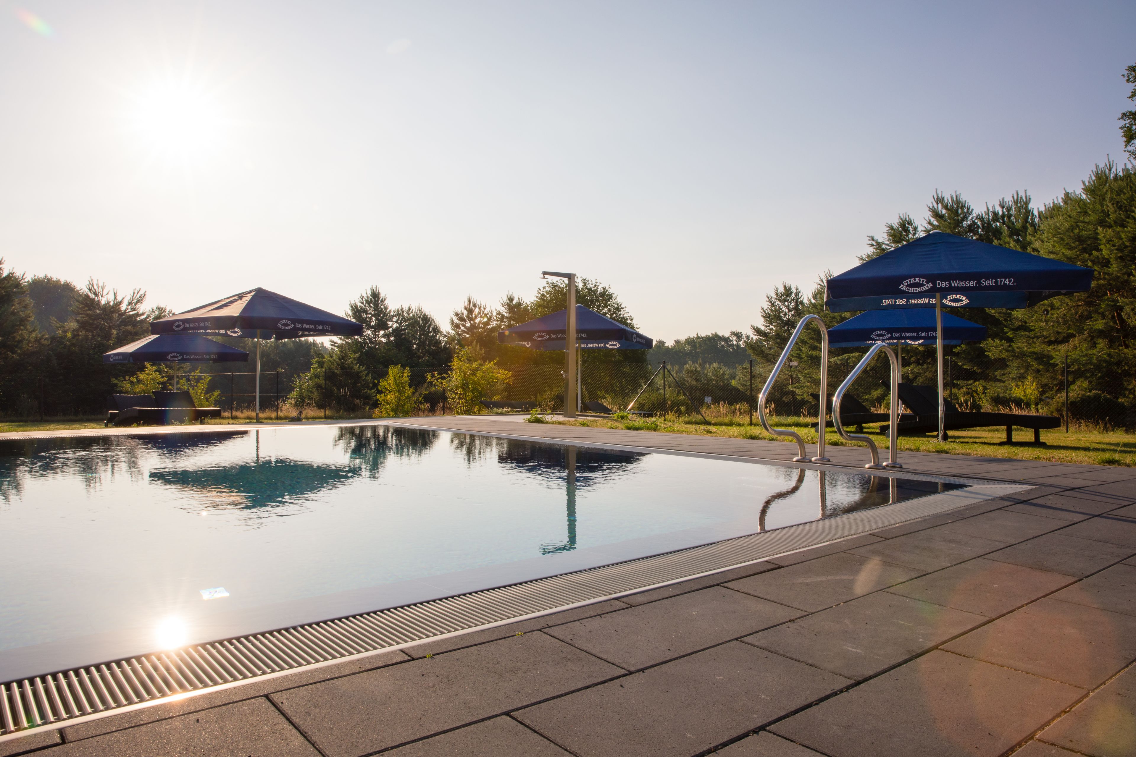 Una piscina all'aperto con acqua cristallina, circondata da un solarium in pietra e ombreggiata da ombrelloni blu.