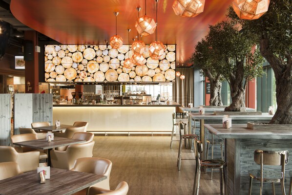 A restaurant with a bar area, wooden tables and chairs, hanging pendant lights, and a large tree in the center. - aja Bad Saarow (Bad Saarow)