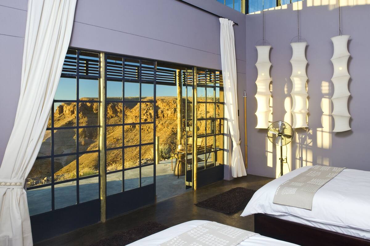 A modern hotel room with a large window offering a view of a desert landscape, a bed with a patterned bedspread, and a unique wall-mounted light fixture.