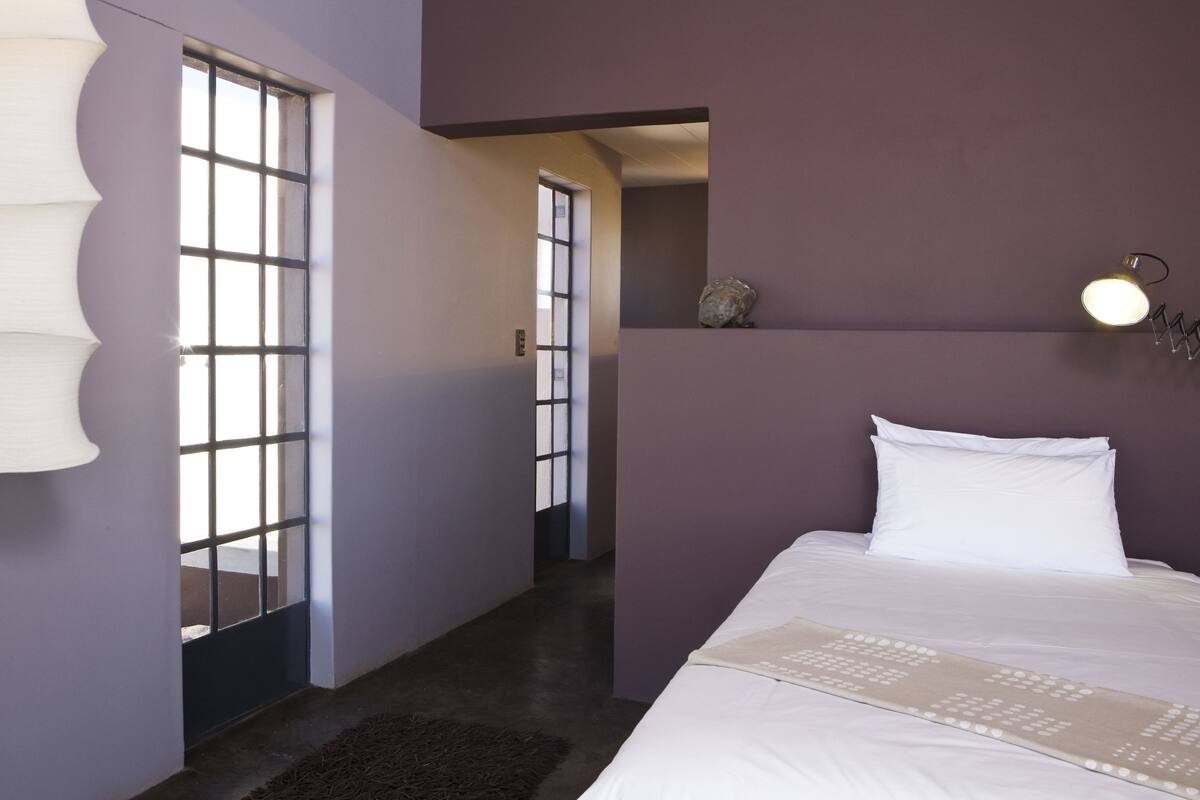 A bedroom with a bed, a nightstand, a wall-mounted lamp, and a window with a view.