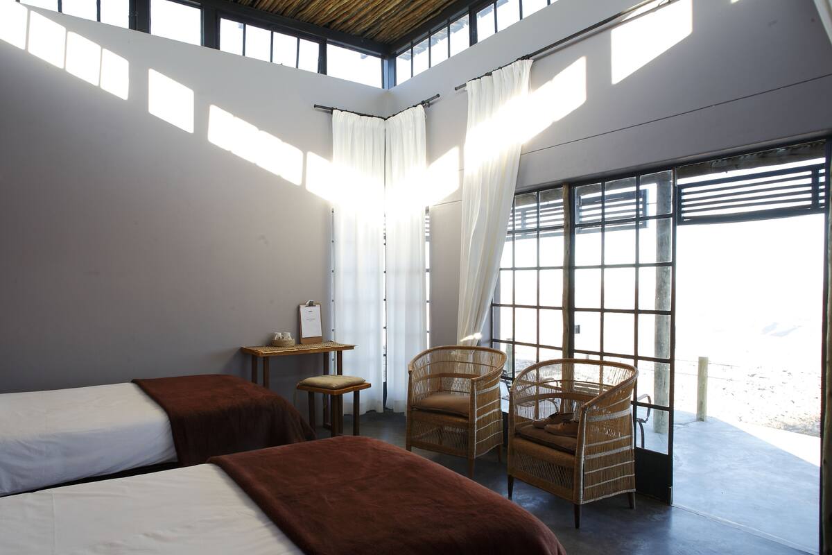 A hotel room with two beds, a small table, a chair, and a large window with curtains.
