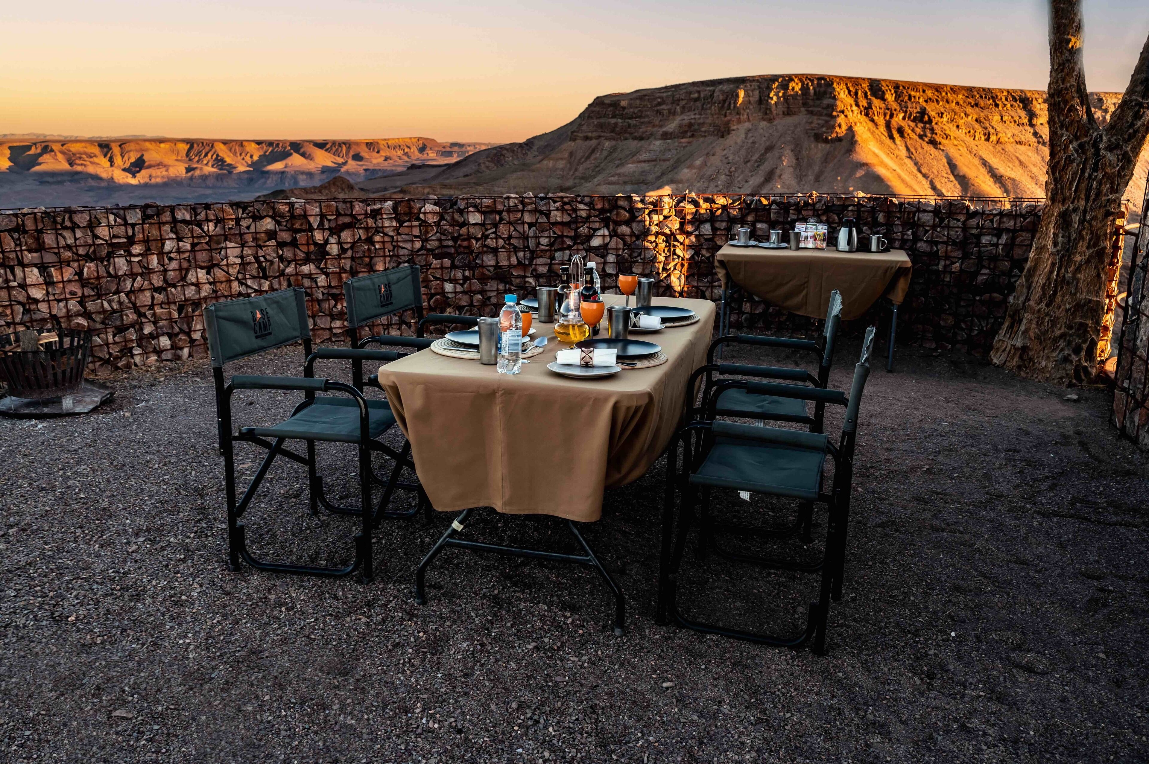Un espacio de comedor al aire libre con una mesa lista para dos personas, rodeada de sillas, frente a un muro de piedra y un paisaje montañoso.
