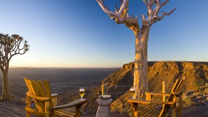 A wooden deck with two chairs and a table, overlooking a scenic landscape with a prominent tree and a view of the horizon. - Fish River Lodge (Fish River Canyon)