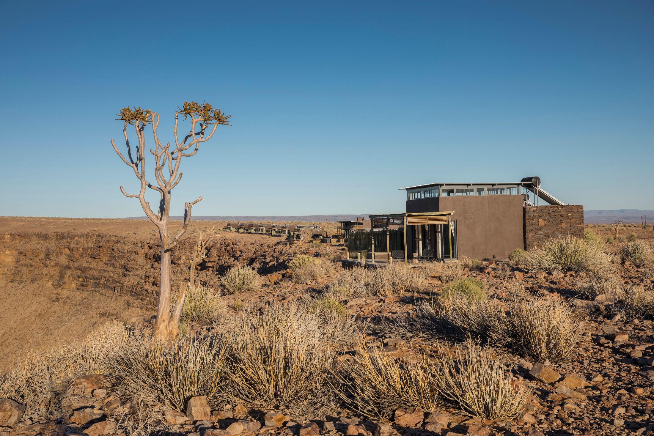 Un edificio moderno con techo plano y un ventanal grande, rodeado de terreno rocoso y árido con vegetación escasa.