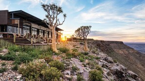 A modern building with a large glass facade, outdoor seating area, and a view of the canyon at sunset. - Fish River Lodge (Fish River Canyon)
