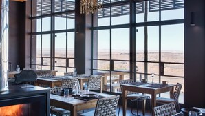 A cozy dining area with a fireplace, wooden furniture, and large windows offering a view of the desert. - Fish River Lodge (Fish River Canyon)