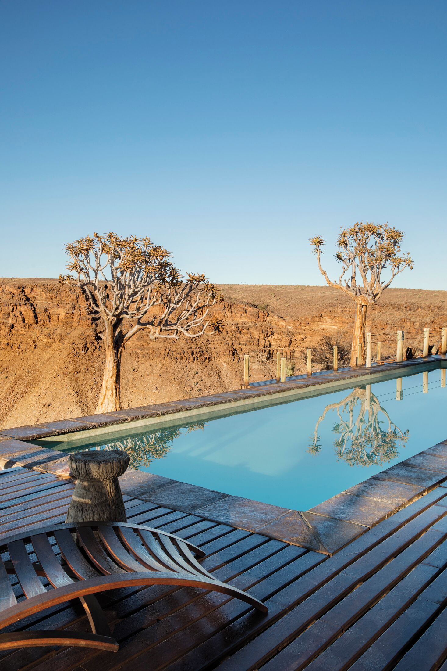 A pool with wooden deck chairs, surrounded by desert landscape and two distinctive trees.