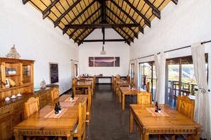 A dining area with wooden tables and chairs, a high ceiling with a wooden beam structure, a chalkboard with writing, and a scenic view through large windows.