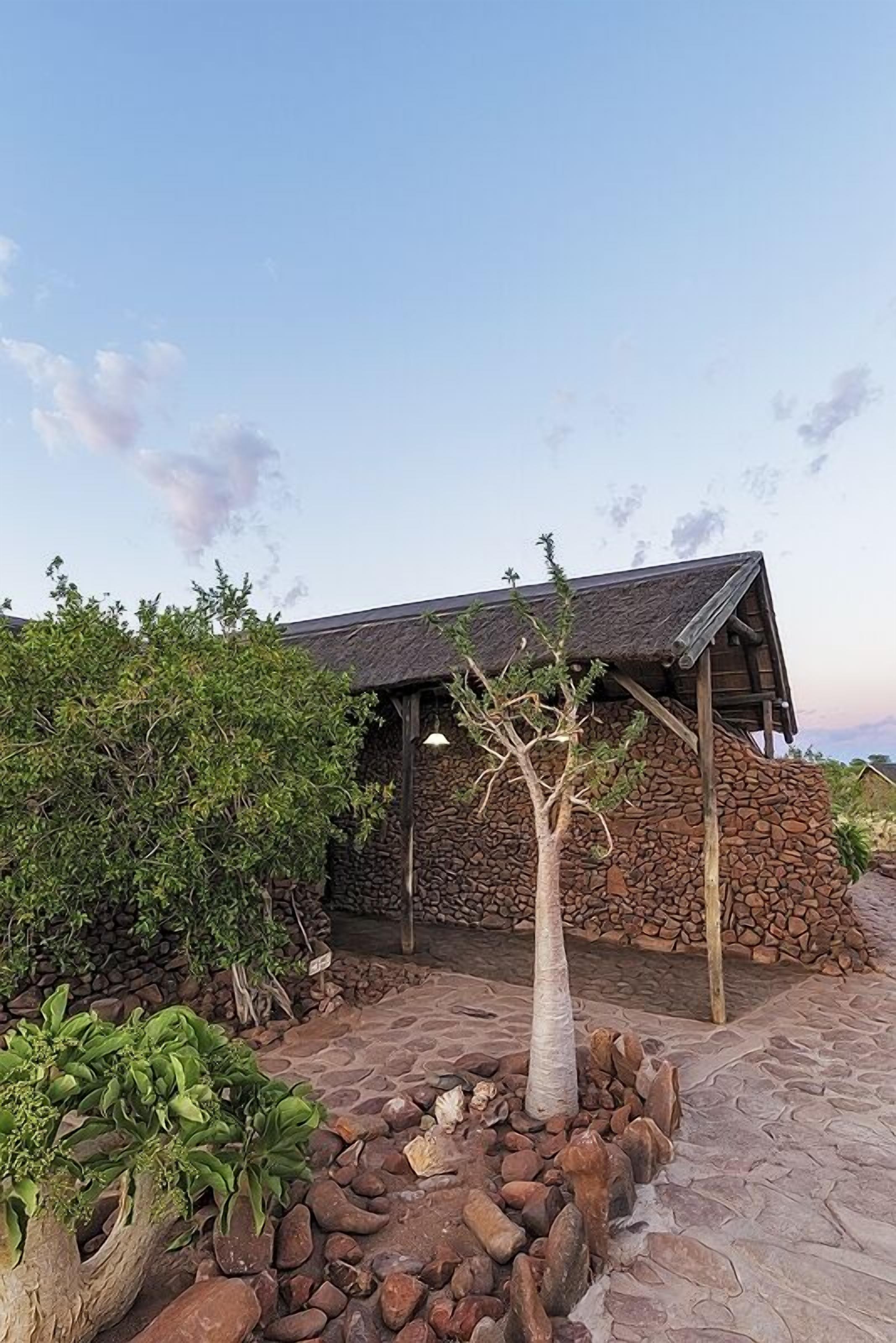 A rustic building with a stone exterior and a thatched roof, surrounded by a landscaped garden with rocks and a small tree.