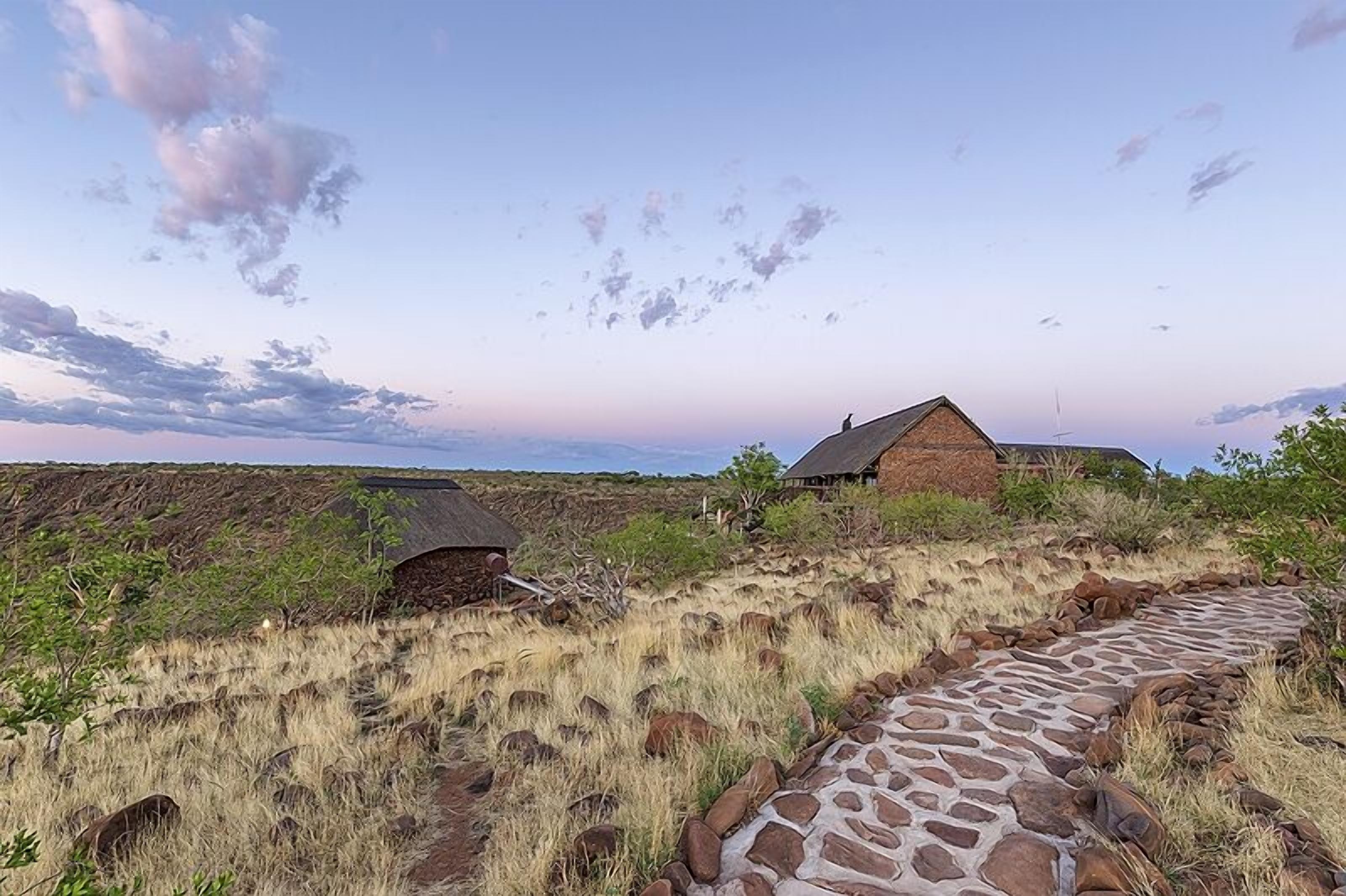 A stone pathway leading to a rustic building with a thatched roof, surrounded by dry grass and sparse vegetation.