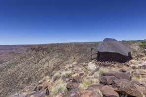 A thatched-roof structure with a staircase leading up to it, situated on a rocky hillside with a clear view of the surrounding landscape.