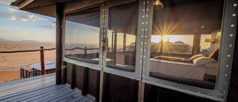 A room with a large window overlooking a desert landscape, a bed with a view, and a wooden deck.