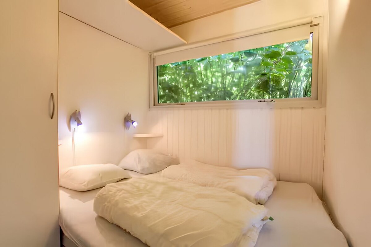 A bedroom with a bed, two bedside lamps, a window with a view of greenery, and a wooden ceiling.