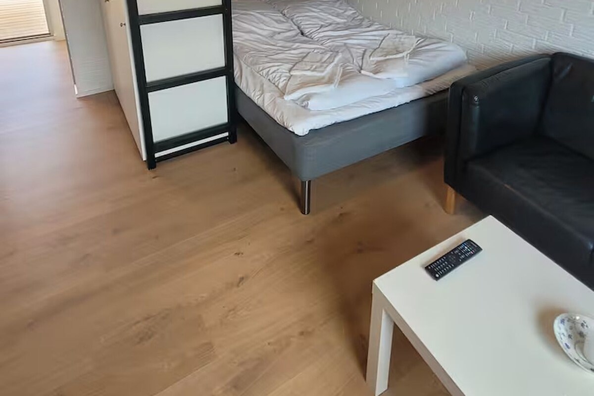 A bunk bed with a ladder, a grey mattress, and a white headboard. There is a black leather sofa and a white table with a remote control and a decorative item.