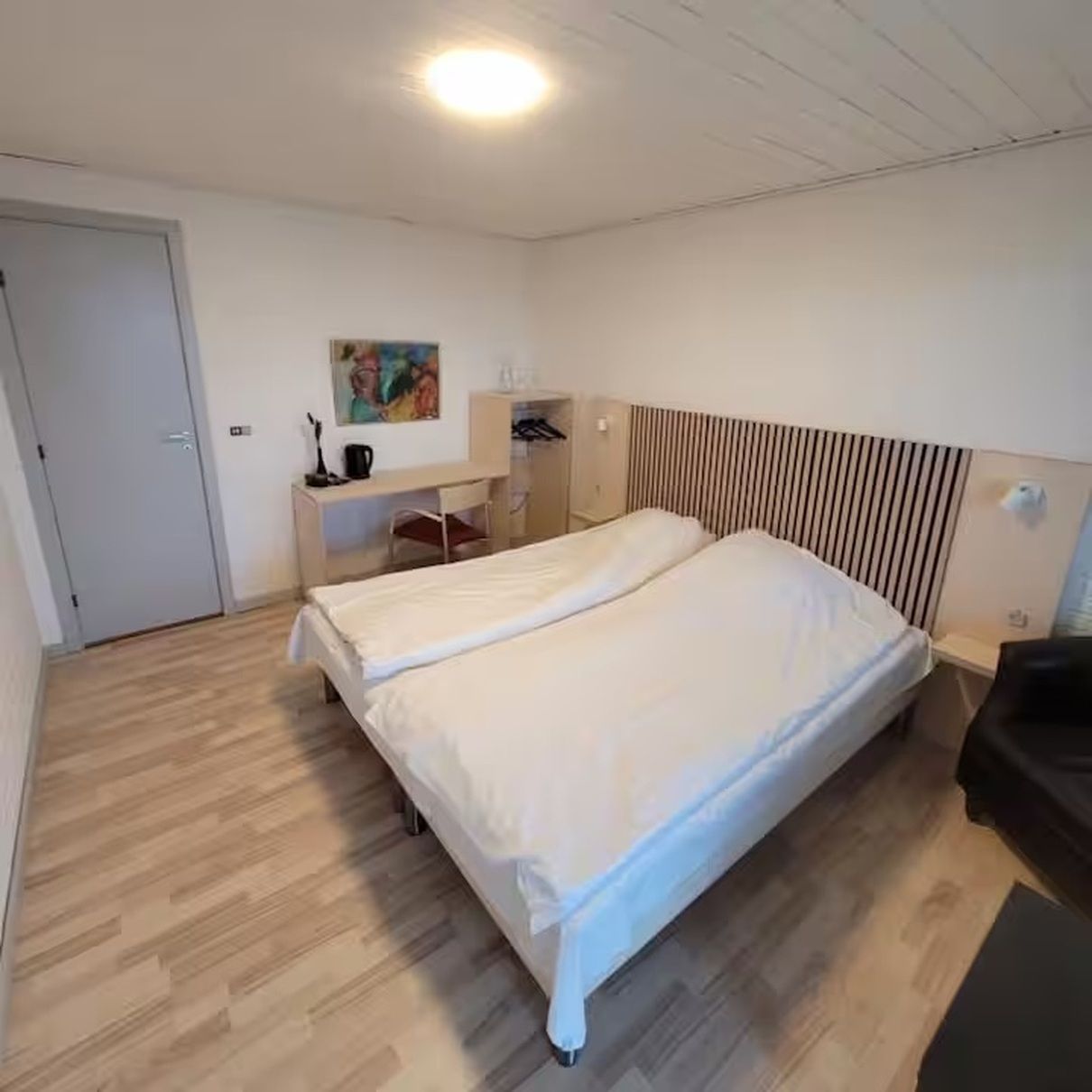 A bedroom with a double bed, a desk with a chair, a wardrobe, and a painting on the wall.