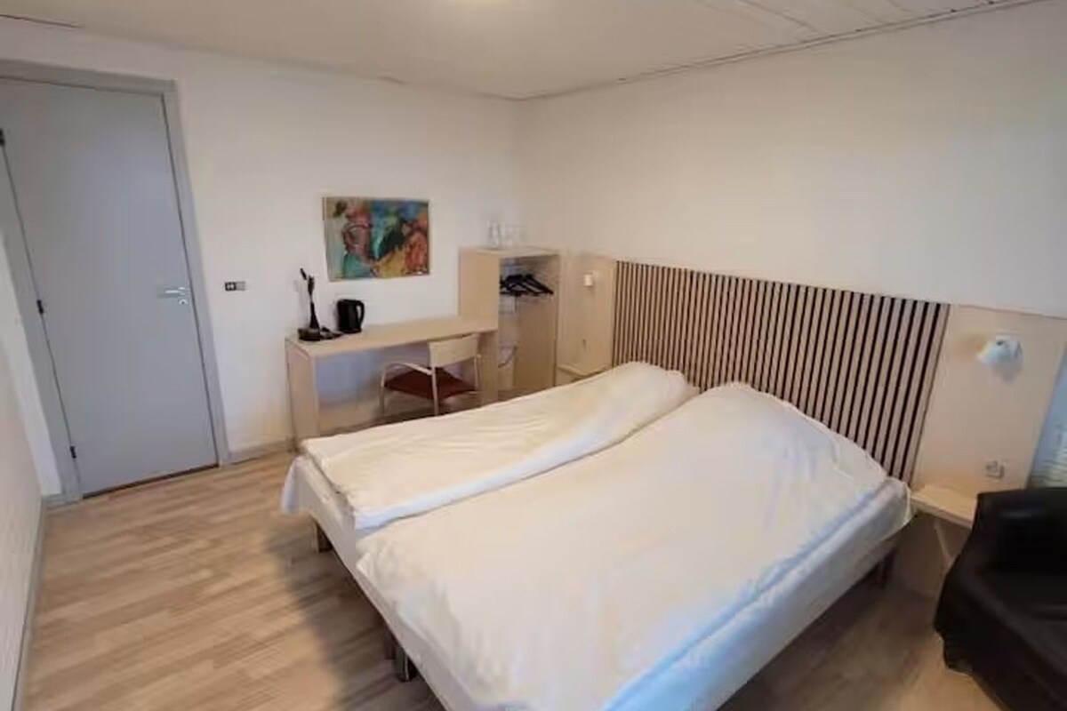 A bedroom with a double bed, a desk with a chair, a wardrobe, and a painting on the wall.