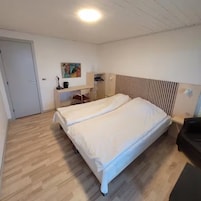 A bedroom with a double bed, a desk with a chair, a wardrobe, and a painting on the wall.