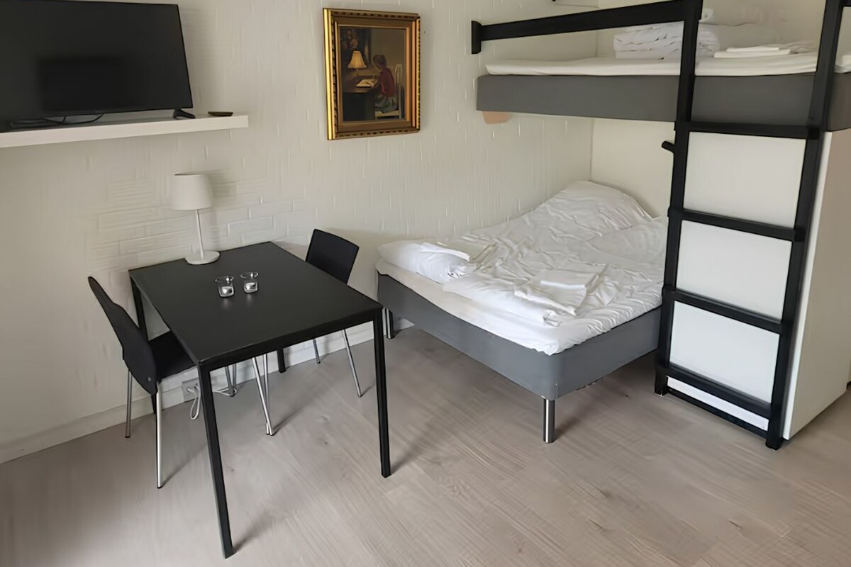 A compact room with a bunk bed, a desk with two chairs, a television on a shelf, and a painting on the wall.