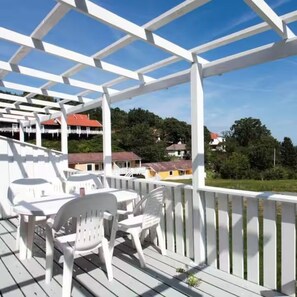 A covered outdoor area with white plastic chairs and tables, a white railing, and a view of houses in the background. - Feriecenter Bornholm ApS (Sandkås)