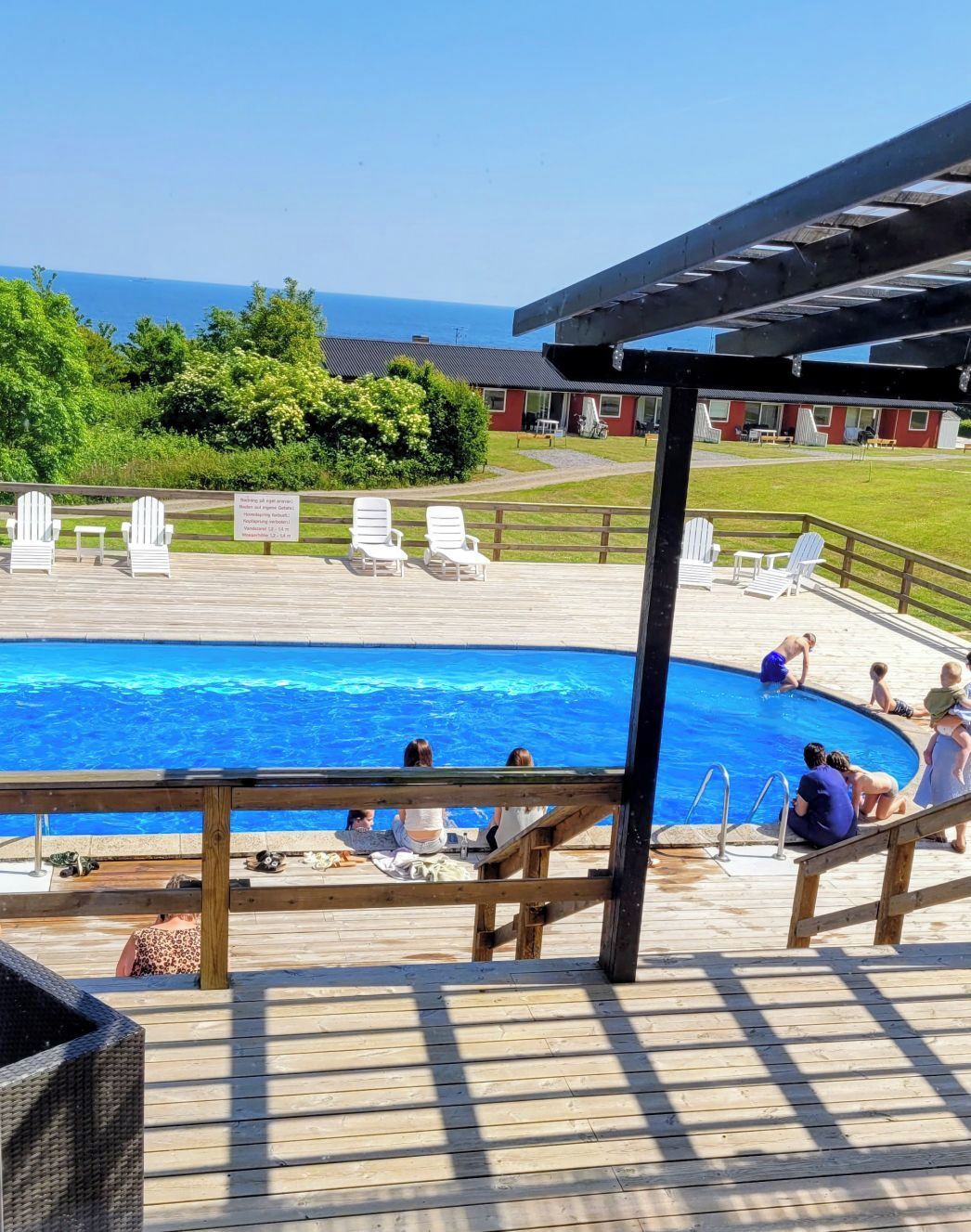 A swimming pool with a wooden deck and lounge chairs, surrounded by a grassy area and a building with a red roof.