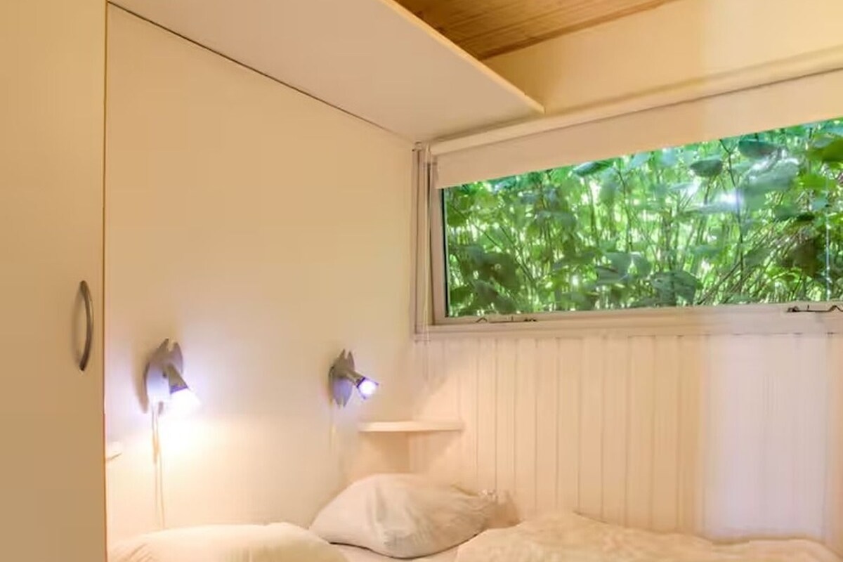 A bedroom with a bed, pillows, a nightstand, and a window with a view of greenery.