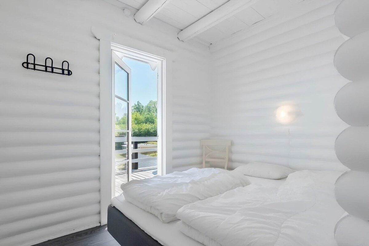 A room with a bed, a small bedside table, a window, and a wall-mounted light fixture.